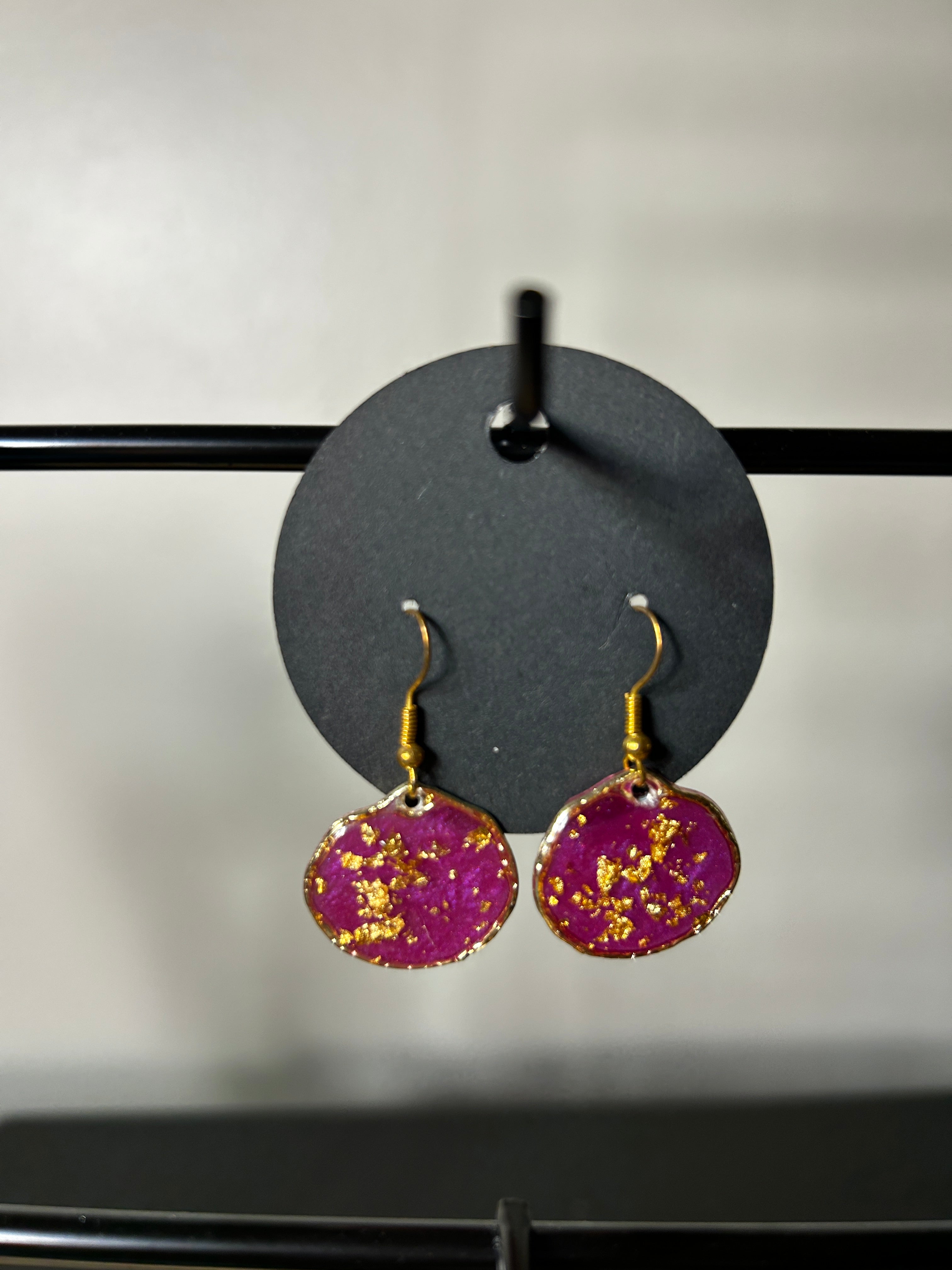 Magenta resin disc drop earrings with gold leaf and gold-tone edge on gold fish hook wires displayed on a dark circular card