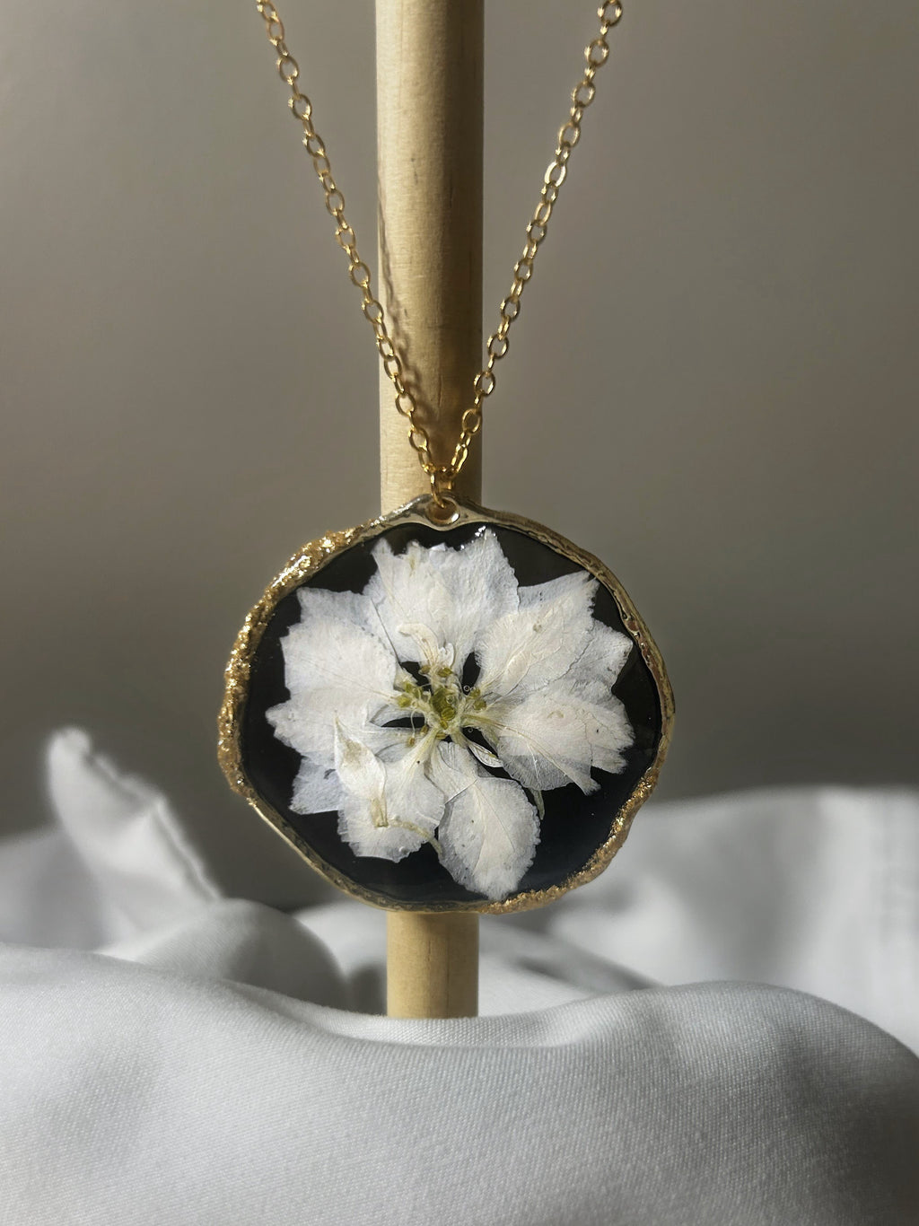 White pressed flower in black resin freeform disc pendant with gold textured edge on delicate gold chain