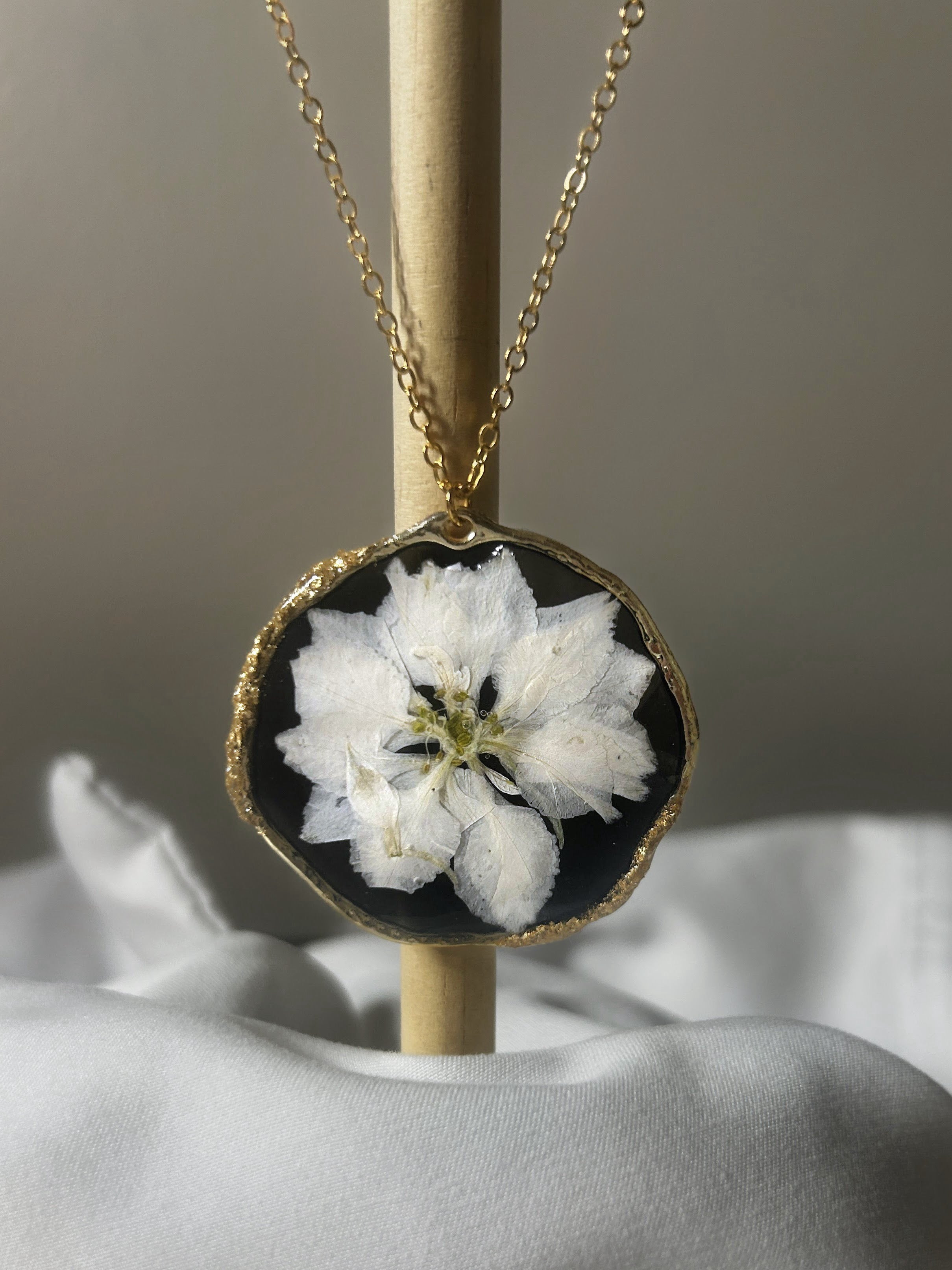 White pressed flower in black resin freeform disc pendant with gold textured edge on delicate gold chain